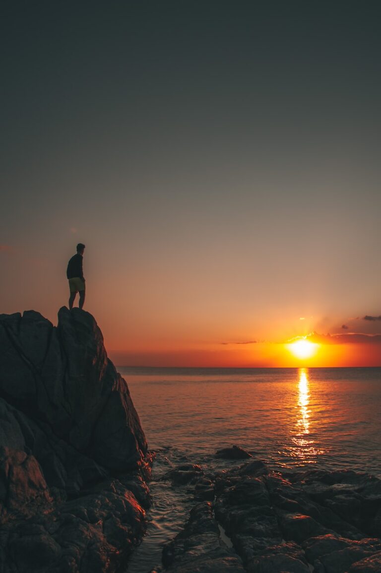 silhouette of person standing on rock formation near body of water during sunset